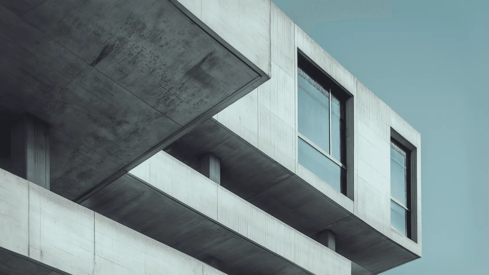 A cantilevered concrete corner with dark windows against a pale blue sky
