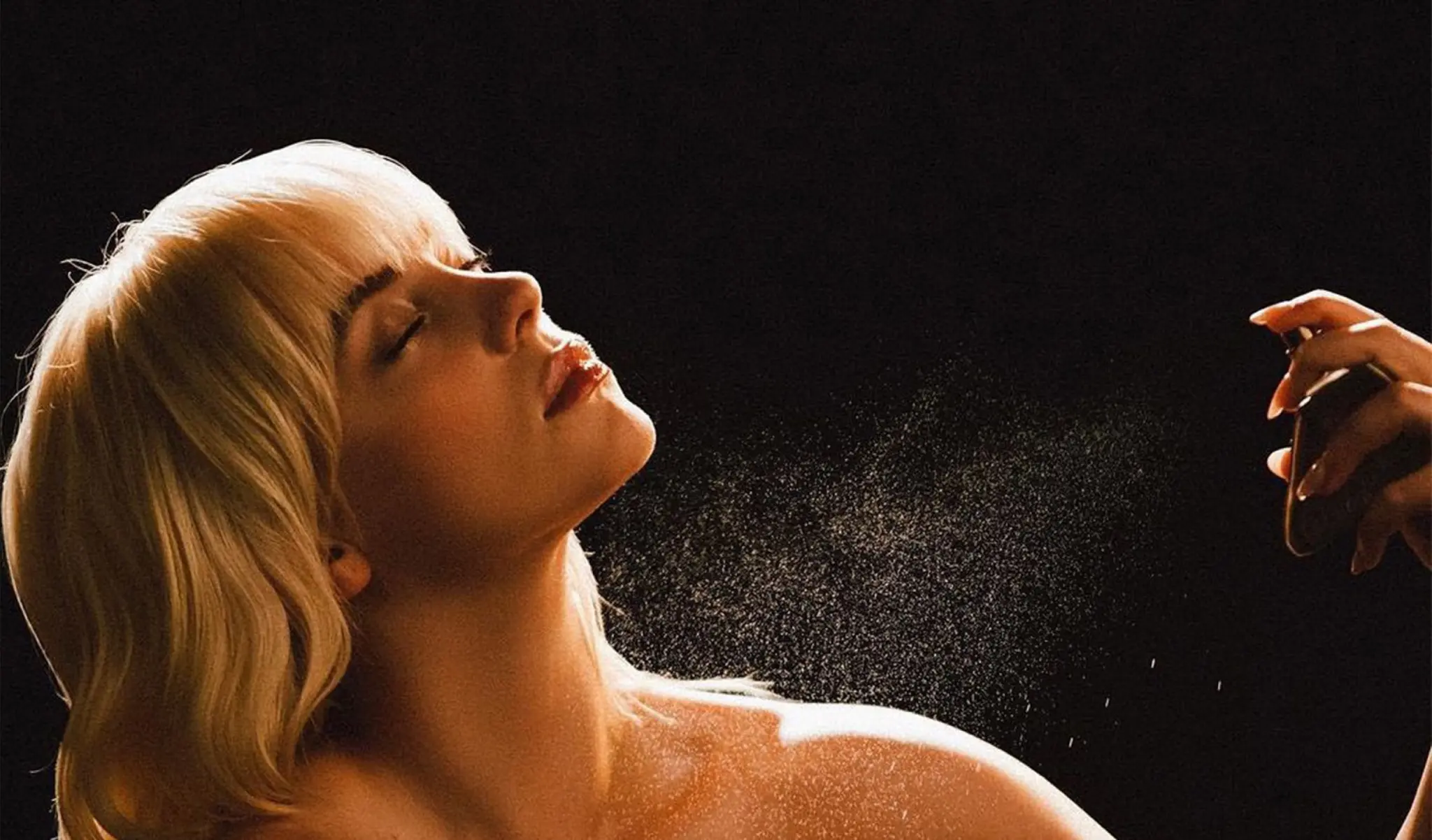 Billie Eilish, eyes closed and head tilted back, as a hand sprays a mist of perfume across her in warm light against black