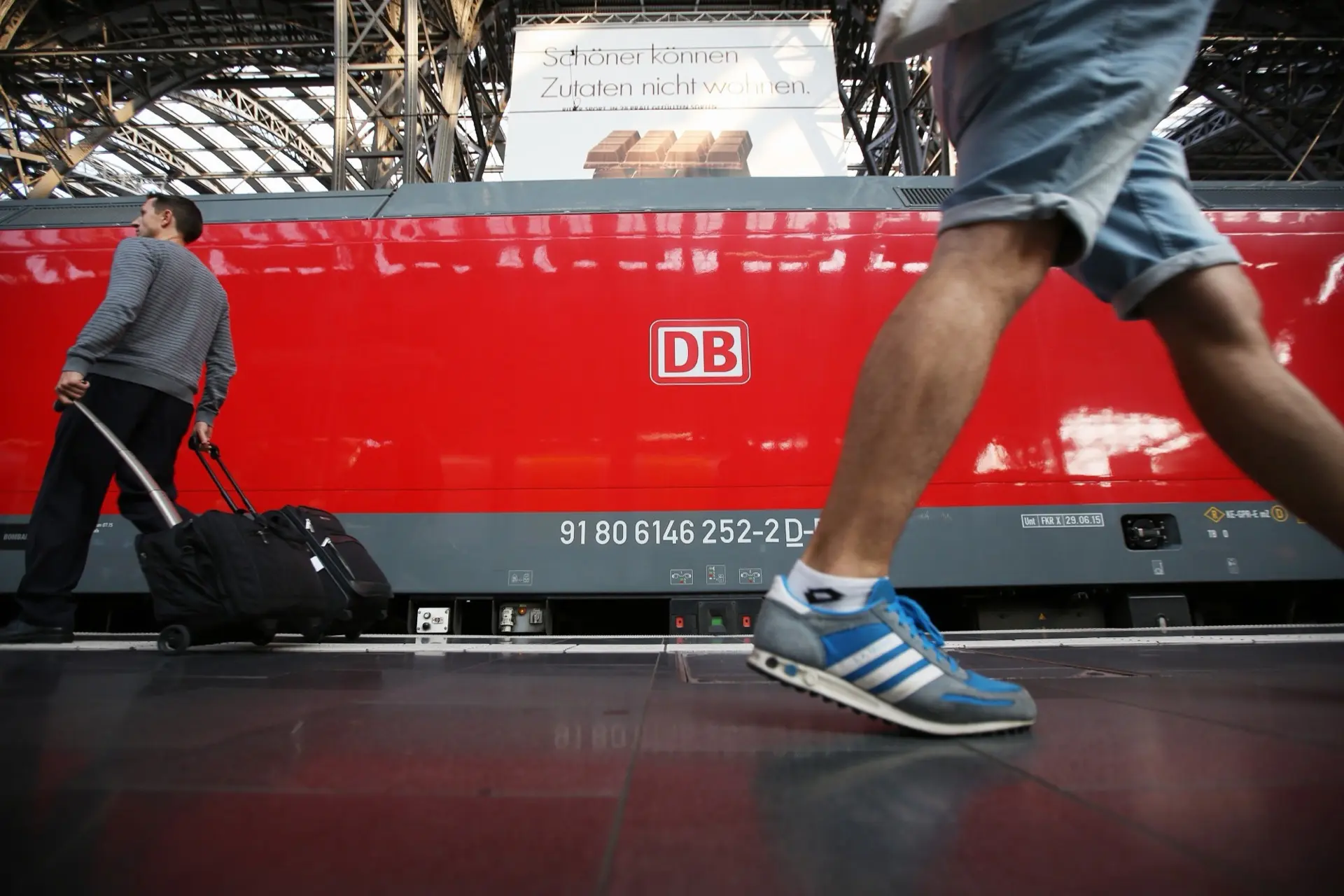 A red Deutsche Bahn locomotive at a station platform, a traveller with a wheeled case beside it and a passer-by striding past in the foreground