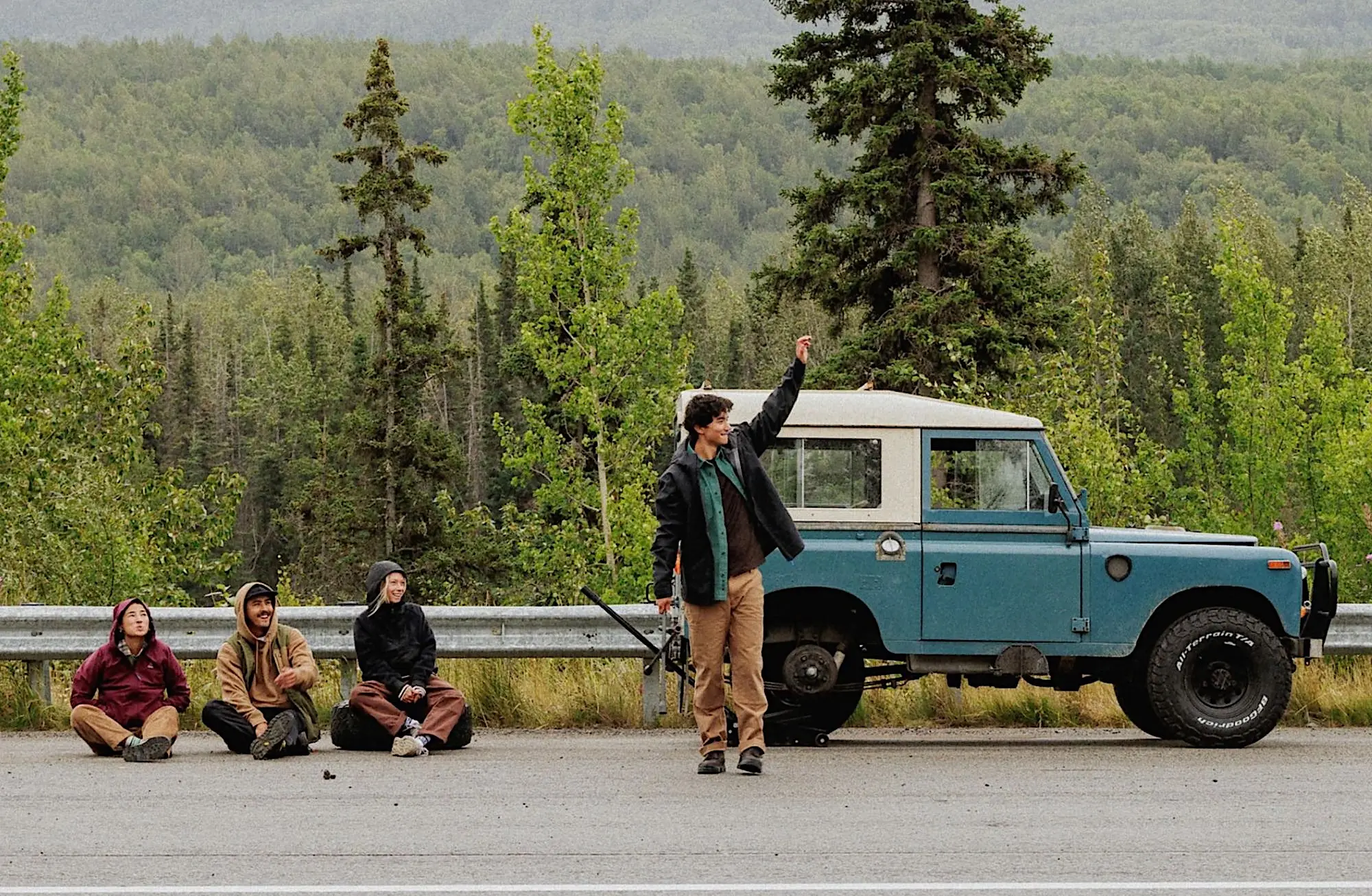 Four friends by a blue Land Rover on a forest highway: three sit on the tarmac while one stands and waves