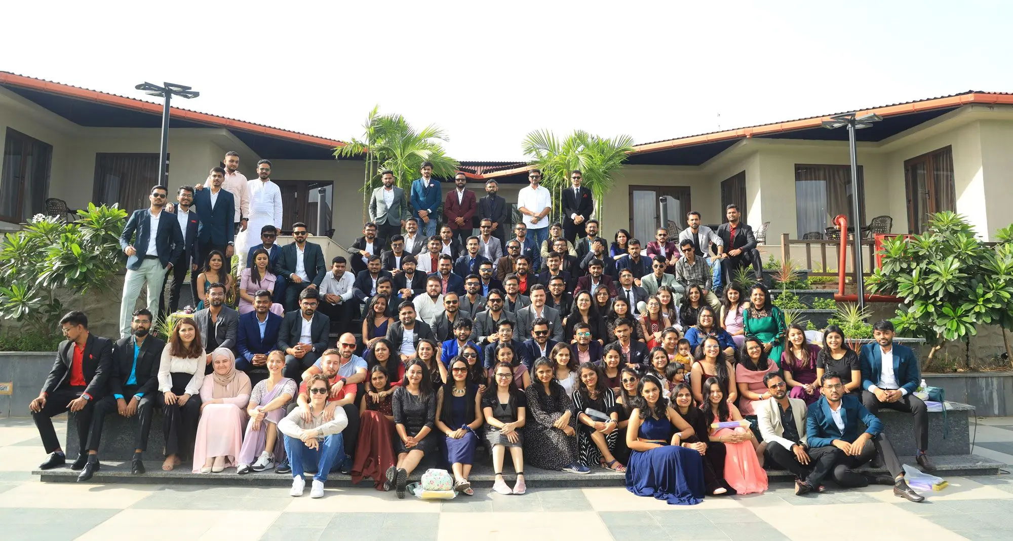 The PlanetX team, about a hundred people dressed up for an event, posed together on the steps of a courtyard in front of a low building with palm trees